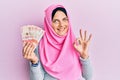 Young caucasian woman wearing islamic hijab holding colombian pesos banknotes doing ok sign with fingers, smiling friendly