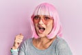 Young caucasian woman wearing pink wig and heart sunglasses pointing thumb up to the side smiling happy with open mouth Royalty Free Stock Photo