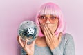 Young caucasian woman wearing pink wig holding disco ball covering mouth with hand, shocked and afraid for mistake Royalty Free Stock Photo