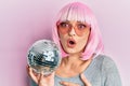 Young caucasian woman wearing pink wig holding disco ball scared and amazed with open mouth for surprise, disbelief face Royalty Free Stock Photo