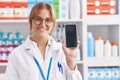 Young caucasian woman working at pharmacy drugstore showing smartphone screen looking positive and happy standing and smiling with Royalty Free Stock Photo