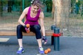Young caucasian woman workouts on the park sports ground, holding dumbbells, sitting on the sport bar; black and lilac sportswear. Royalty Free Stock Photo