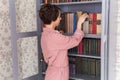 Young caucasian young woman with brunette hair taking boor for reading on free time standing near bookcase, positive