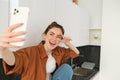 Young charismatic girl takes selfies in the kitchen, posing for photo, holds mobile phone with extended hand