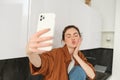 Young charismatic girl takes selfies in the kitchen, posing for photo, holds mobile phone with extended hand