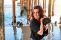 A young charming brunette woman in a black shirt and denim shorts poses under a pier on the beach of Malibu, California Royalty Free Stock Photo