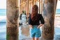 A young charming brunette woman in a black shirt and denim shorts poses under a pier on the beach of Malibu, California Royalty Free Stock Photo