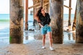 A young charming brunette woman in a black shirt and denim shorts poses under a pier on the beach of Malibu, California Royalty Free Stock Photo