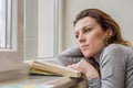 Young charming girl student, with long hair, sad at the window reading book with lessons Royalty Free Stock Photo