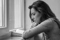 Young charming girl student, with long hair, sad at the window reading book with lessons Royalty Free Stock Photo