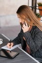 Young charming woman is calling on a mobile phone, sitting in a cafe and talking on a mobile phone with a smile, writing notes in Royalty Free Stock Photo