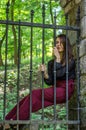 Young charming woman with long hair offender, sits behind bars in an ancient stone castle fortress prison prisoner and looks Royalty Free Stock Photo