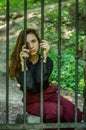 Young charming woman with long hair offender, sits behind bars in the old stone jail prisoner and looks pityingly through steel ba