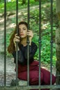 Young charming woman with long hair offender, sits behind bars in the old stone jail prisoner and looks pityingly through steel ba