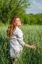 Young charming woman walking outdoors in a field near the green bushes and trees, hand patting wheat ears, dressed in a beautiful Royalty Free Stock Photo