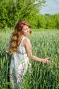 Young charming woman walking outdoors in a field near the green bushes and trees, hand patting wheat ears, dressed in a beautiful Royalty Free Stock Photo