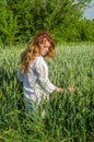 Young charming woman walking outdoors in a field near the green bushes and trees, hand patting wheat ears, dressed in a beautiful Royalty Free Stock Photo