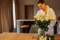 Young charming woman in white bathrobe cuts stems of white roses and puts them in glass vase Royalty Free Stock Photo