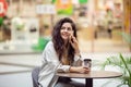 Young cheerfful woman using smartphone and drinking take away coffee in paper cup. Stylish woman sitting at cafe indoors Royalty Free Stock Photo