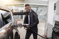 Young cheerful African American businessman refueling his modern luxury car at fuel station, smiling to camera