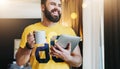 Young bearded man stands in room and holding tablet computer while drinking coffee. Guy freelancer working at home. Royalty Free Stock Photo