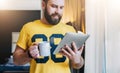 Cheerful bearded man stands and using tablet computer. Guy laughs looking screen of digital tablet while drinking coffee Royalty Free Stock Photo