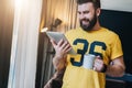 Cheerful bearded man stands and using tablet computer. Guy laughs looking screen of digital tablet while drinking coffee Royalty Free Stock Photo