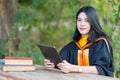 A young cheerful beautiful female gradute student wearing academic gown studying in college campus with books and lablet on table Royalty Free Stock Photo