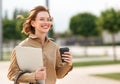 Young cheerful businesswoman walks outside with beaming smile while holding cup of coffee and laptop Royalty Free Stock Photo