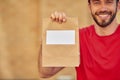 Young cheerful caucasian male courier in red uniform holding small paper bag with copy space for text and smiling at Royalty Free Stock Photo