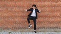 Young cheerful caucasian man in a hat and black suit dances against a brick wall and takes off his hat, copy space, slow