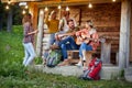 Young cheerful friends toasting and drinking beer in front of wooden cottage on the terrace. Girl playing guitar. Summertime Royalty Free Stock Photo