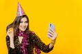 Young cheerful girl 20s in a festive hat and New Year`s tinsel takes a selfie or broadcasts live on a smartphone Royalty Free Stock Photo