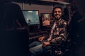 A young cheerful Indian guy wearing a military shirt sitting on a gamer chair and looking at a camera in a gaming club