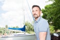 Young cheerful man relaxing on sailboat posing and looking at far on background of boats, sky and trees. Royalty Free Stock Photo