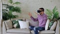 Young cheerful man wearing virtual reality headset having 360 VR video experience while sitting on couch in living room Royalty Free Stock Photo