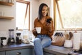 Young cheerful mixed race woman drinking a cup of coffee while using her phone alone in the morning in the kitchen. One Royalty Free Stock Photo