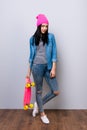 Young cheerful stylish hipster girl in pink hat and jeans holding skateboard in her hand against gray background Royalty Free Stock Photo