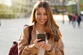 Young cheerful stylish woman using cell phone and texting message on city street. Beautiful happy casual girl holding smart phone Royalty Free Stock Photo