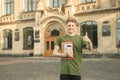 Young cheerful successful caucasian nerdy student is showing thumb up, standing with books, university on background, near campus