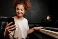 Young cheerful woman happily using phone playing on electric piano in recording studio