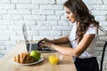 Young cheerful woman using on laptop computer and eating fruits in the kitchen in the morning Royalty Free Stock Photo