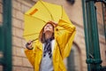 A young cheerful woman with a yellow raincoat and umbrella who is enjoying while listening to the music and walking the city on a Royalty Free Stock Photo