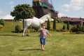 A young child boy of 3 years in a striped T-shirt barefoot on the grass goes to a white horse. Summer sunny childhood