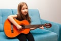 Young child, little school age girl playing classic acoustic guitar, practicing musical instrument sitting on the sofa