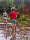 Young children in rubber boots pull toys on a rope along a country sandy road with puddles after rain