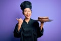 Young chinese chef woman wearing cooker uniform and hat holding tray with cake pointing and showing with thumb up to the side with Royalty Free Stock Photo