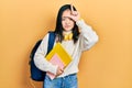 Young chinese girl holding student backpack and books making fun of people with fingers on forehead doing loser gesture mocking