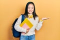 Young chinese girl holding student backpack and books smiling cheerful presenting and pointing with palm of hand looking at the Royalty Free Stock Photo