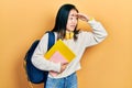 Young chinese girl holding student backpack and books very happy and smiling looking far away with hand over head